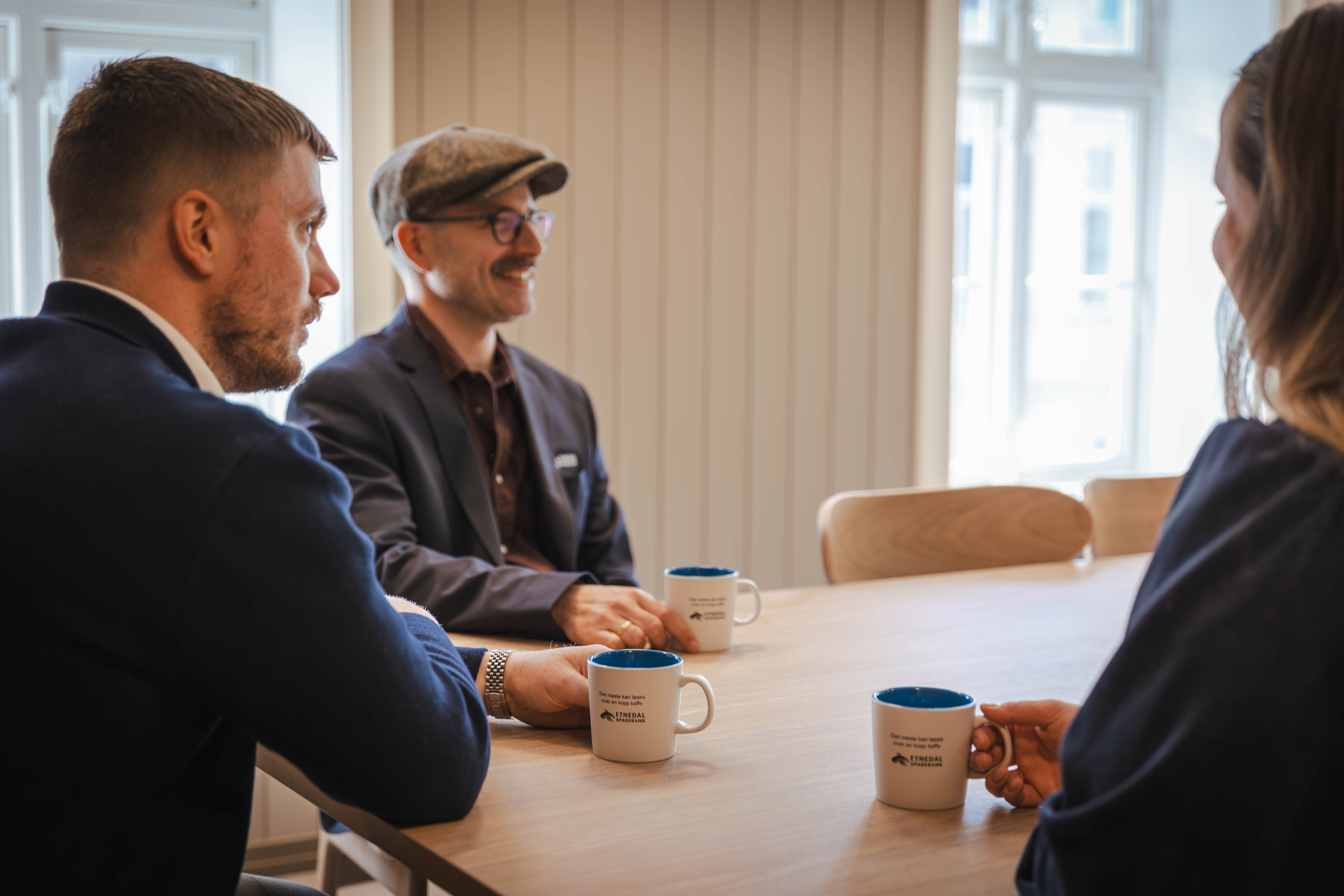 Tre personer, to menn og en dame, sitter ved et bord med kaffekopper i hendene og snakker sammen. 