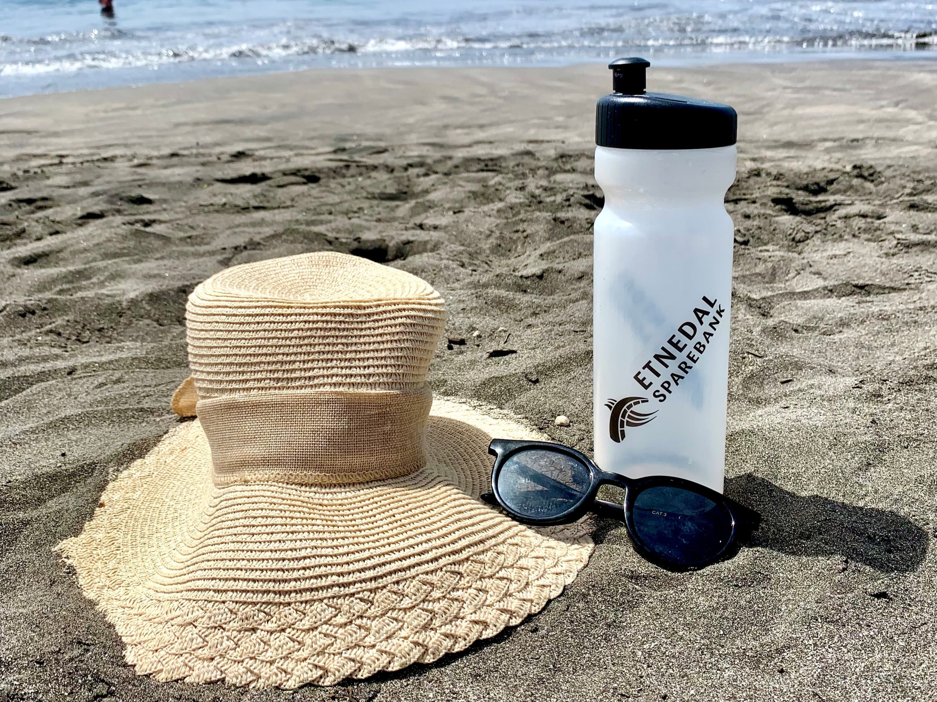 Ståhatt, solbriller og flaske med Etnedal Sparebanks logo på en strand