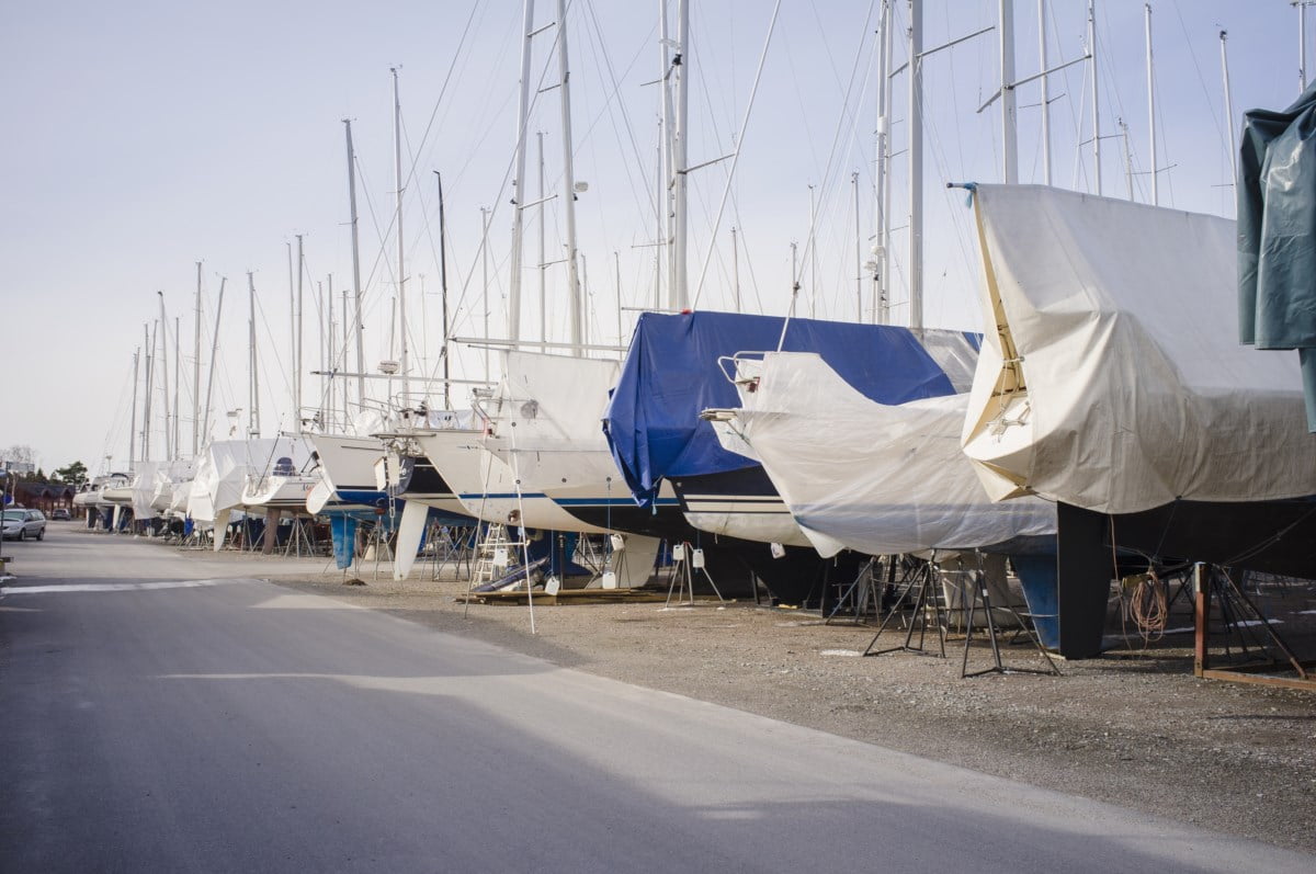 Seilbåter på land dekket med presenninger for vinteropplag, linet opp i en marina med master som stikker opp i bakgrunnen mot en klar himmel.
