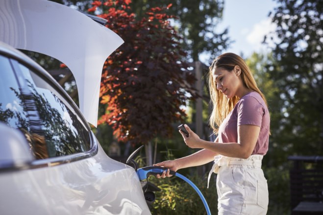 Ung kvinne lader elbilen sin og ser på mobilen når hun gjør det.